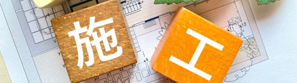 建築図面の上に木製ブロックで「施工」と書かれた文字が置かれているイメージ。住宅施工や工事に関する内容を連想させる構成。