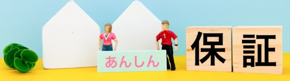 木製ブロックに「保証」、プレートに「あんしん」と書かれた文字と住宅の模型、ミニチュアの男女が並ぶ、安心感を伝えるイメージ。