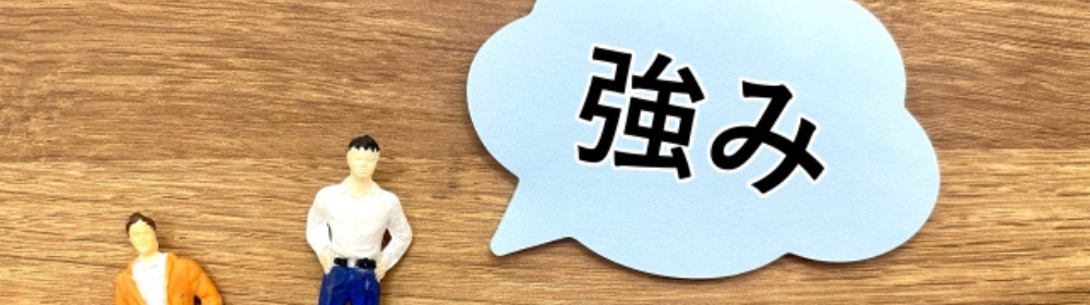 木目の背景にミニチュアの人物2体と、「強み」と書かれた吹き出しプレートが置かれている構図。